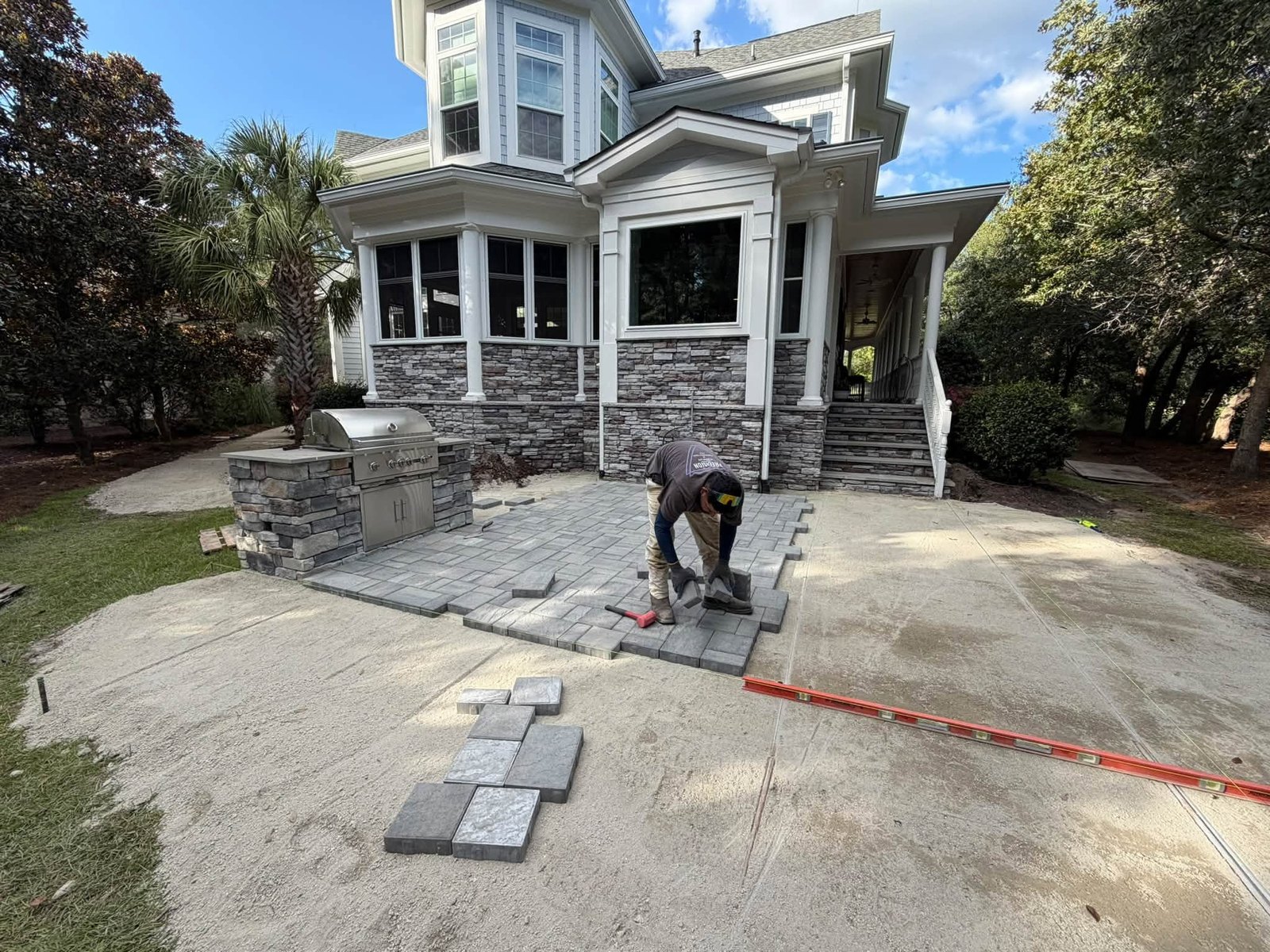 Paver patio installation in progress behind a coastal home in Ocean Ridge Plantation, with a Precision Hardscape crew member setting pavers next to a stone outdoor kitchen